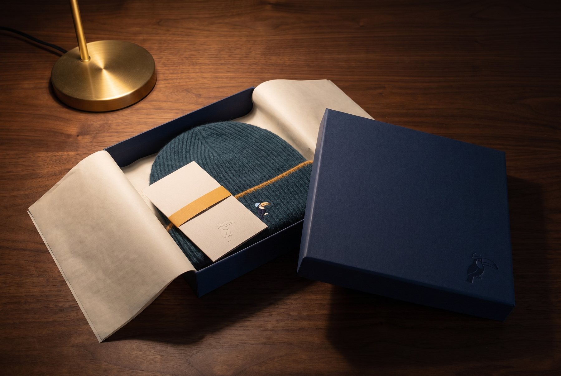 A night-blue rigid box open on a walnut table under a brass lamp: bone tissue folded back from a petrol hat, a bone card with an ochre band, the bird blind-embossed on the lid.