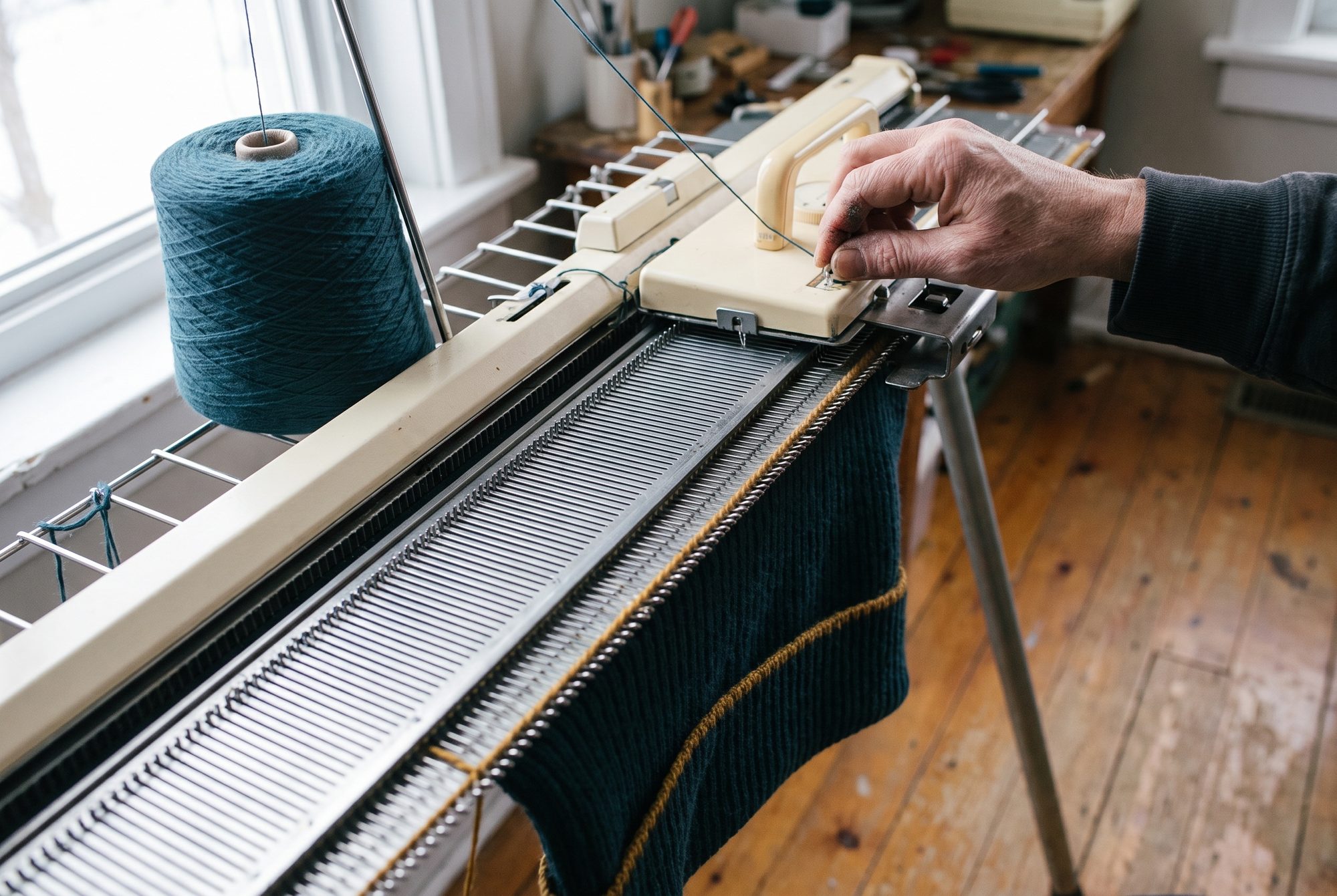 The needle bed of a flat-bed knitting machine knitting petrol rib with one ochre line, a cone of yarn beside, grey winter light.