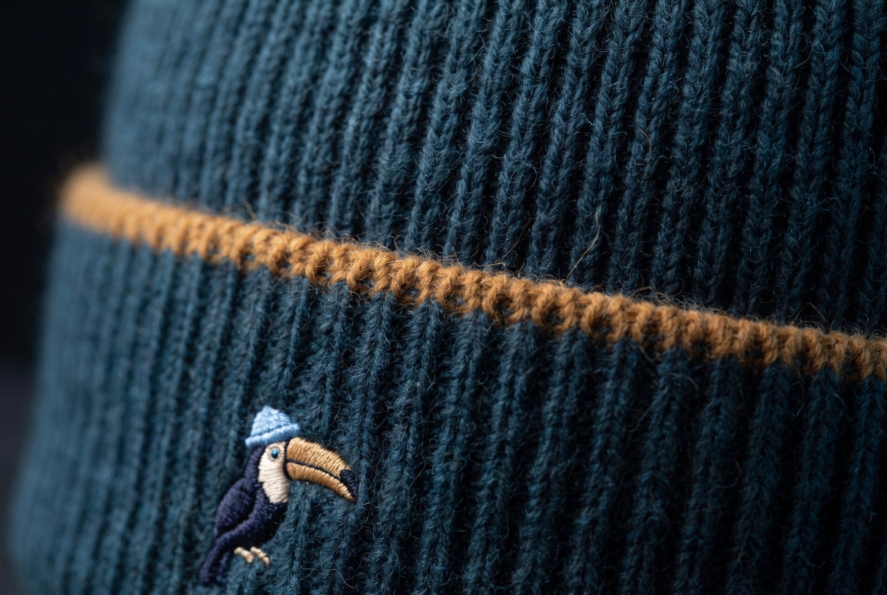 Close view of the Watch: the rib of petrol cashmere, the single ochre line at the fold of the cuff, and the small embroidered toucan in its ice-blue cap.